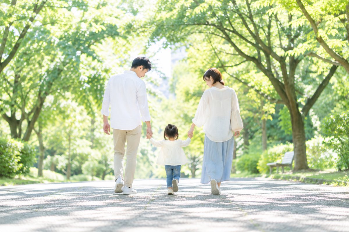 公園の木立の中を、手をつないで歩く両親と子どもの後ろ姿